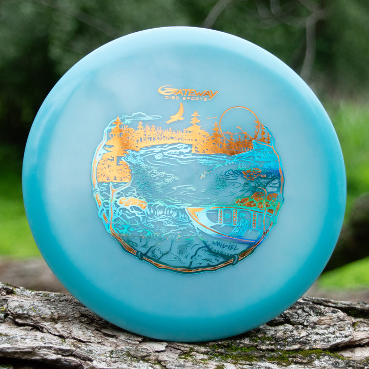 Sam Smigiel's Element - NXT Color Superglow disc golf disc, a blue mid-range with gold and blue nature scene design and reliable straight flight, stands upright on wood outdoors.