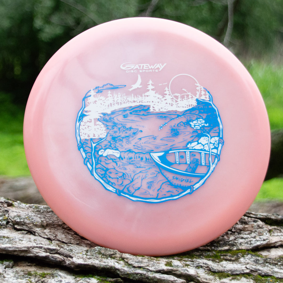 A pink Sam Smigiel's Element - NXT Color Superglow disc golf disc with great glide, featuring a blue and white river scene with a canoe, trees, and rocks, displayed outdoors on a tree trunk.