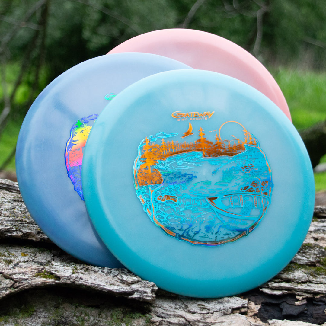 Three vibrant Sam Smigiel's Element - NXT Color Superglow discs with detailed landscape designs rest on a log, offering exceptional glide for straight flights through lush green foliage in the background.