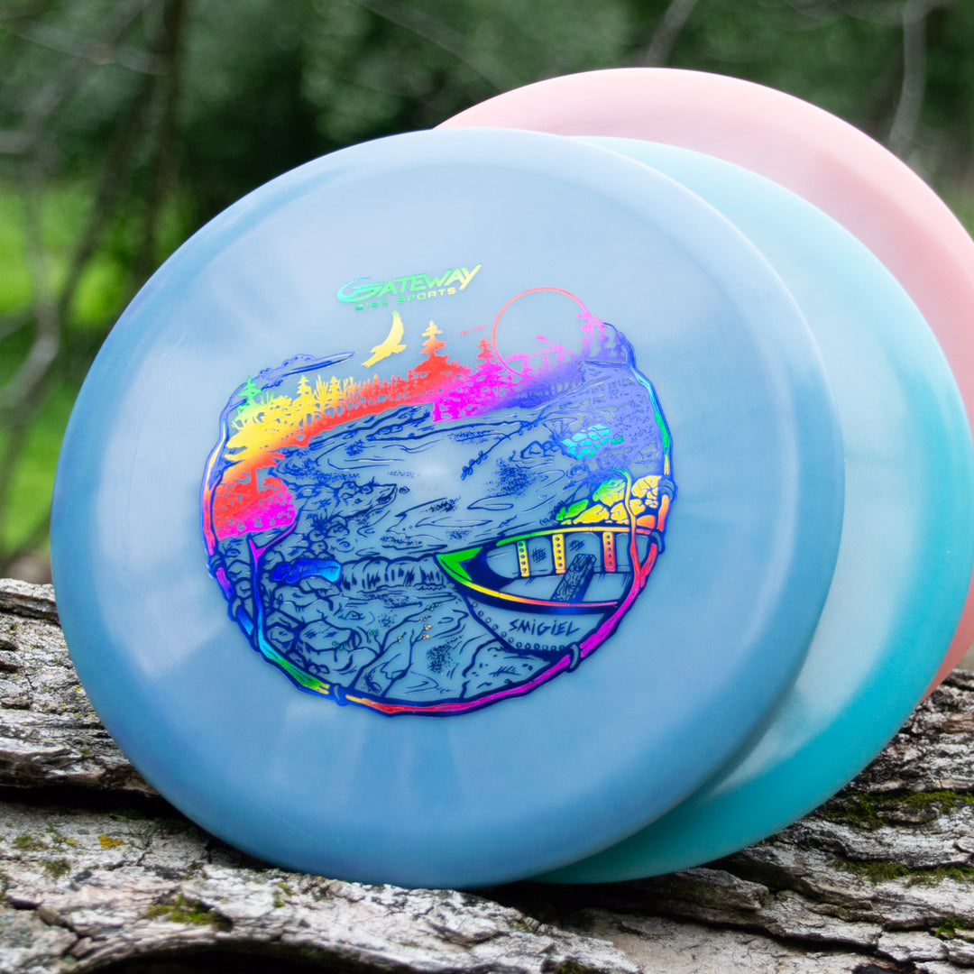 A close-up of two stacked disc golf discs on a log, with the top one—a Sam Smigiel's Element - NXT Color Superglow mid-range—showing a vibrant landscape graphic and Gateway Disc Sports logo, designed for straight flight and amazing glide.