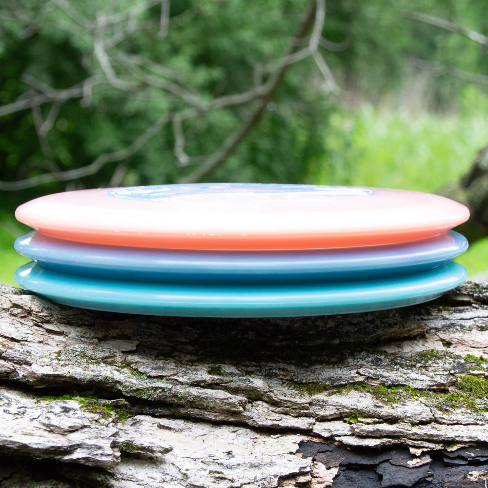 Three NXT Color Superglow discs from Sam Smigiel's Element, in soft pastel hues, are stacked on a textured log, showcasing incredible glide and straight flight against a misty green forest backdrop.
