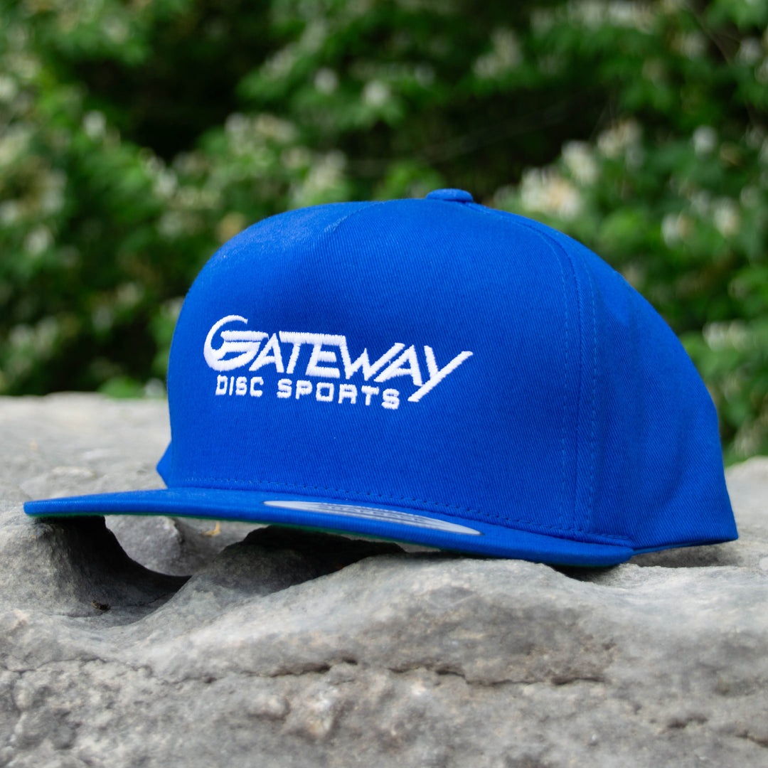A Gateway Logo'd SnapBack Hat, featuring "Gateway Disc Sports" embroidered in white on blue, rests on a gray rock with green foliage in the background.