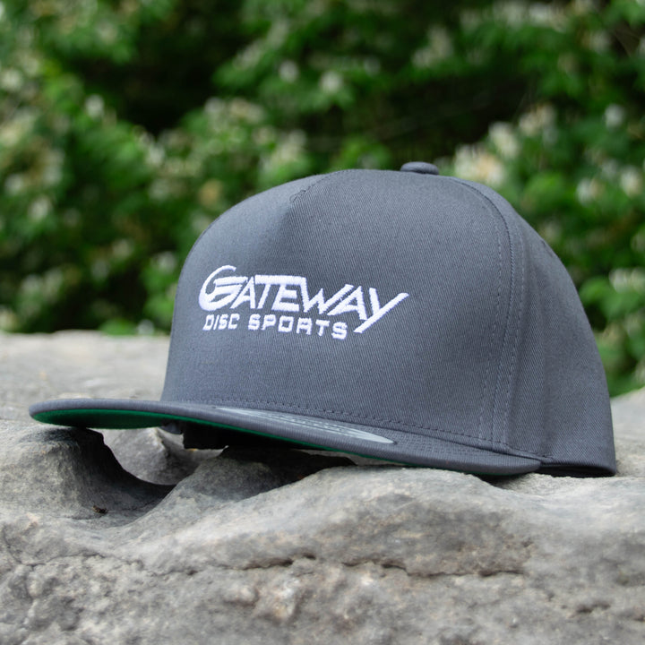A Gateway Logo'd SnapBack Hat in gray, featuring white embroidered lettering, rests on a rock with blurred green foliage in the background.