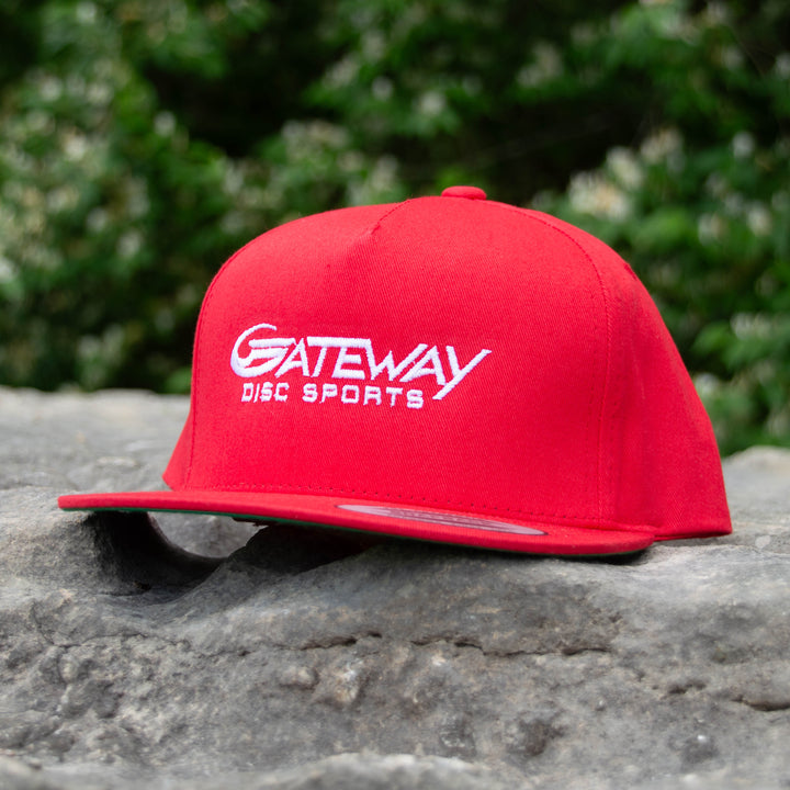The Gateway Logo'd SnapBack Hat, a red five-panel cotton twill cap with white "Gateway Disc Sports" embroidery, sits on a gray rock with greenery in the background.