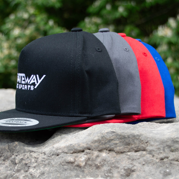 Gateway Logo'd SnapBack Hats in black, gray, red, and blue cotton twill are lined up on stone with greenery softly blurred in the background.