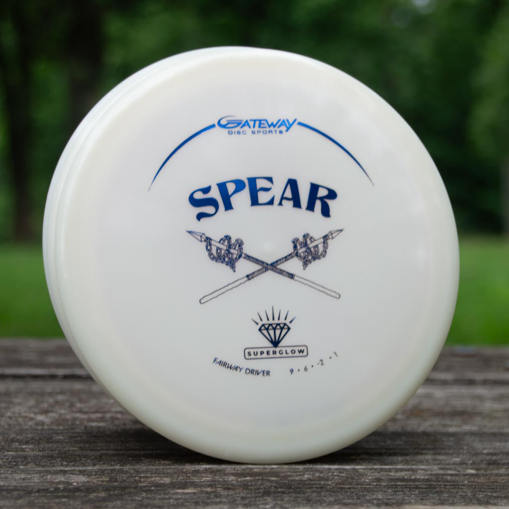 A white Spear - Diamond Superglow fairway driver from Gateway Disc Sports stands upright on a wooden surface, ready for your next throw, with grassy trees in the background.