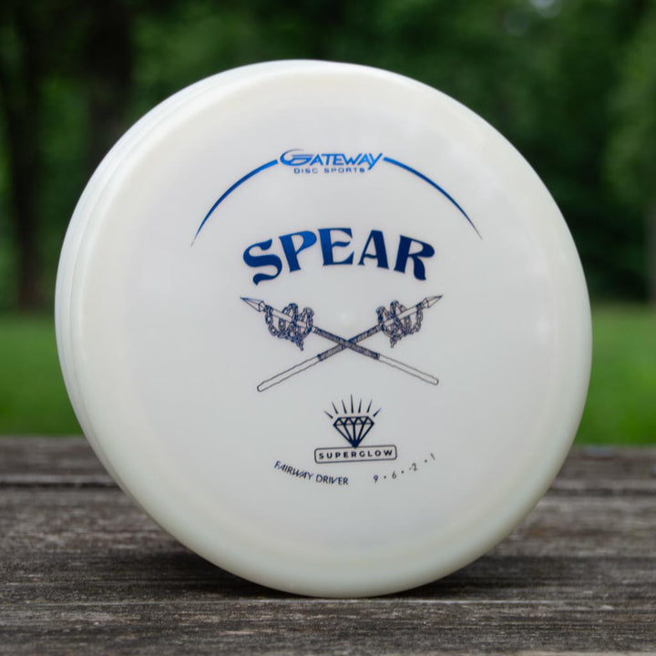 A white Spear - Diamond Superglow fairway driver from Gateway Disc Sports stands upright on a wooden surface, ready for your next throw, with grassy trees in the background.