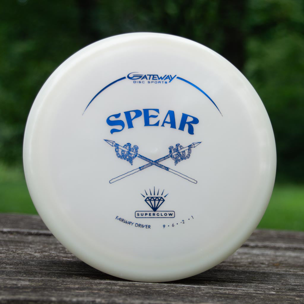 A white "Spear - Diamond Superglow" disc golf driver by Gateway Disc Sports, with two crossed spears, blue text, and flight numbers, lies on a wooden surface outdoors.