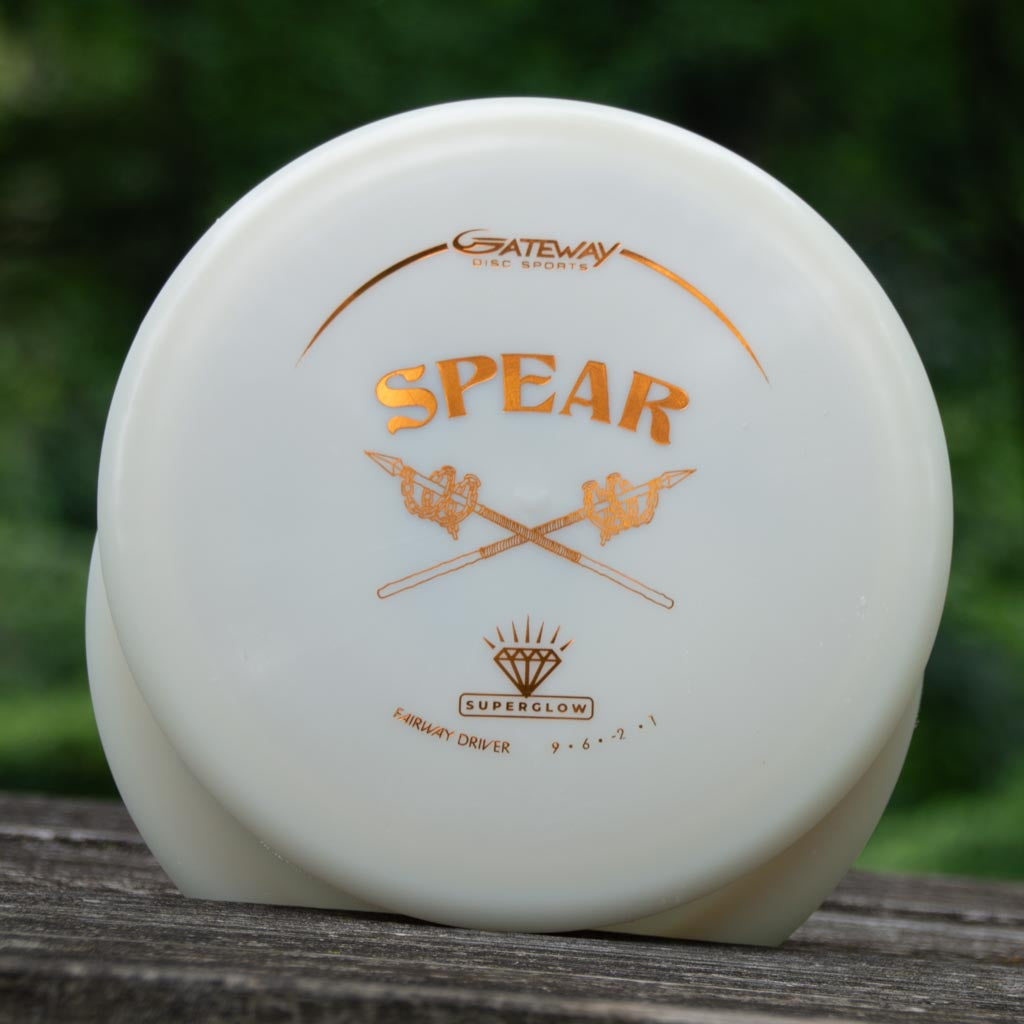 A white disc golf driver labeled "Spear - Diamond Superglow" with gold text, a crossed spears design, and its Flight Numbers sits on a wooden surface with a blurred green background.