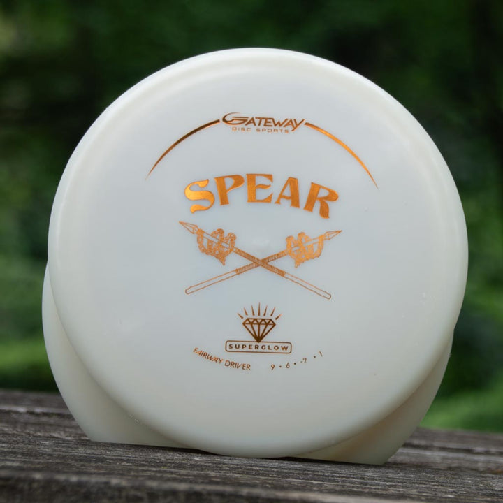 A white disc golf driver labeled "Spear - Diamond Superglow" with gold text, a crossed spears design, and its Flight Numbers sits on a wooden surface with a blurred green background.