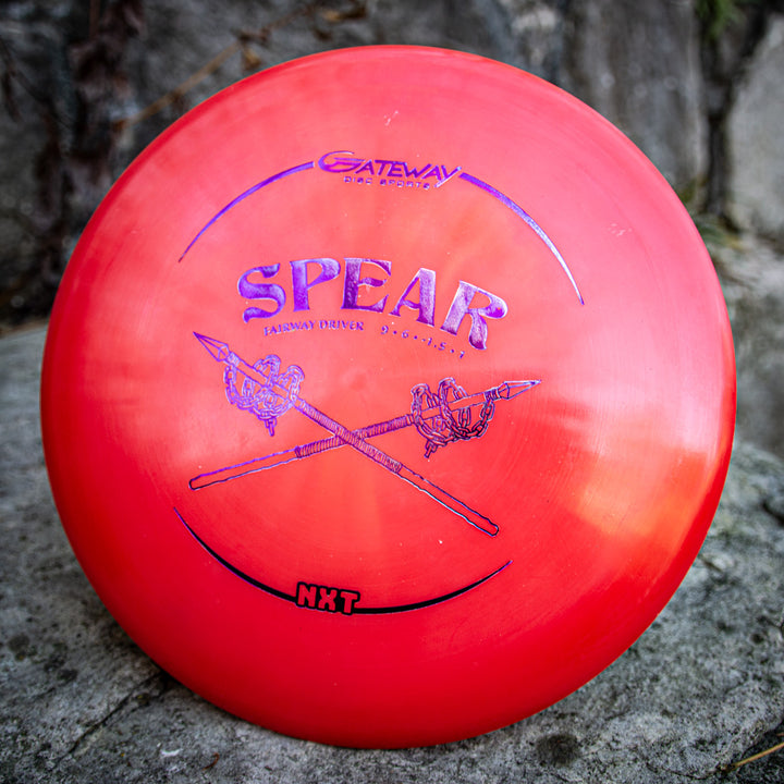 A red Spear - NXT straight control driver with blue crossed spears rests on a rock. Made from a durable plastic blend, it's designed to be your longest flying disc on the course.