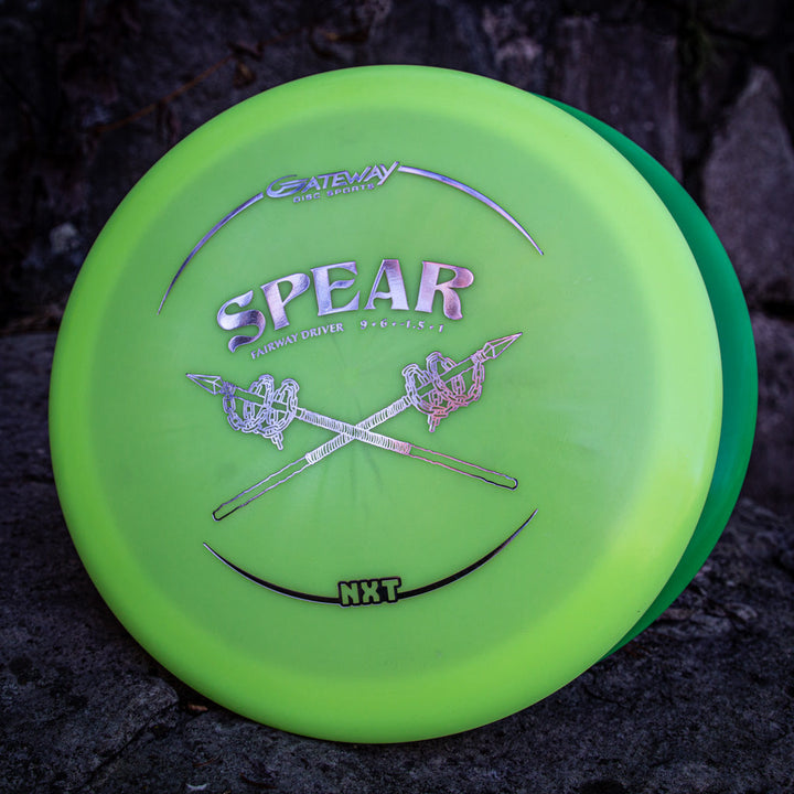 A green Spear - NXT fairway driver disc golf disc, made from a durable plastic blend, featuring a crossed spears graphic and "NXT" text, rests on a stone surface.