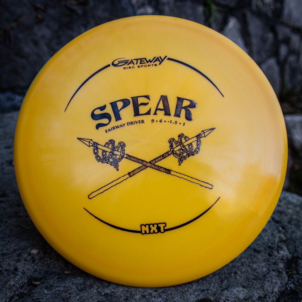 A yellow Spear - NXT straight control driver by Gateway Disc Sports, made from durable plastic blend, features crossed spears graphics and flight numbers on the front, displayed on a rock surface.