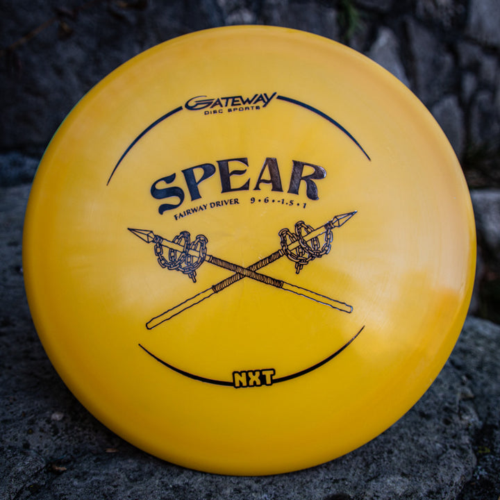 A yellow Spear - NXT straight control driver by Gateway Disc Sports, made from durable plastic blend, features crossed spears graphics and flight numbers on the front, displayed on a rock surface.