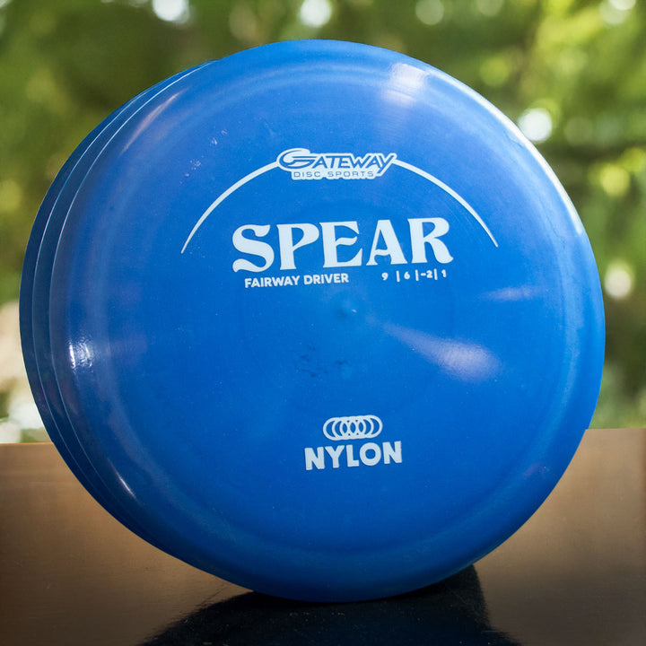 A blue Spear - Nylon control driver disc golf disc with white text rests on a dark surface, highlighting its straight flight potential against a backdrop of blurred green foliage.