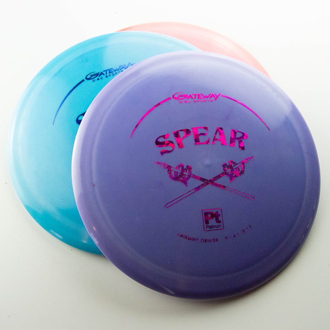Two plastic disc golf discs—a purple "Spear - Platinum" control driver with crossed spears graphic, partially overlapping a blue disc—are shown on a white background. Built for accurate placement and reliable performance.