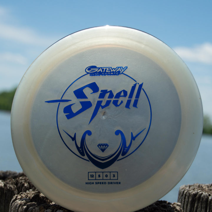 A translucent blue overstable driver labeled "Spell - Diamond," made from high-grade TPU, stands upright on a weathered wooden post beside the water.