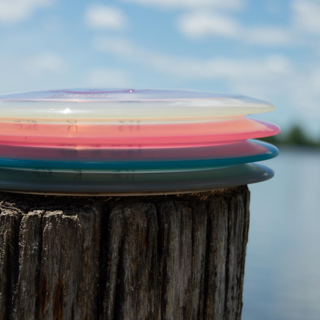 The Spell - Diamond, an overstable disc golf driver, sits atop a weathered wooden post with three colorful discs, set against a lake and cloudy sky backdrop.