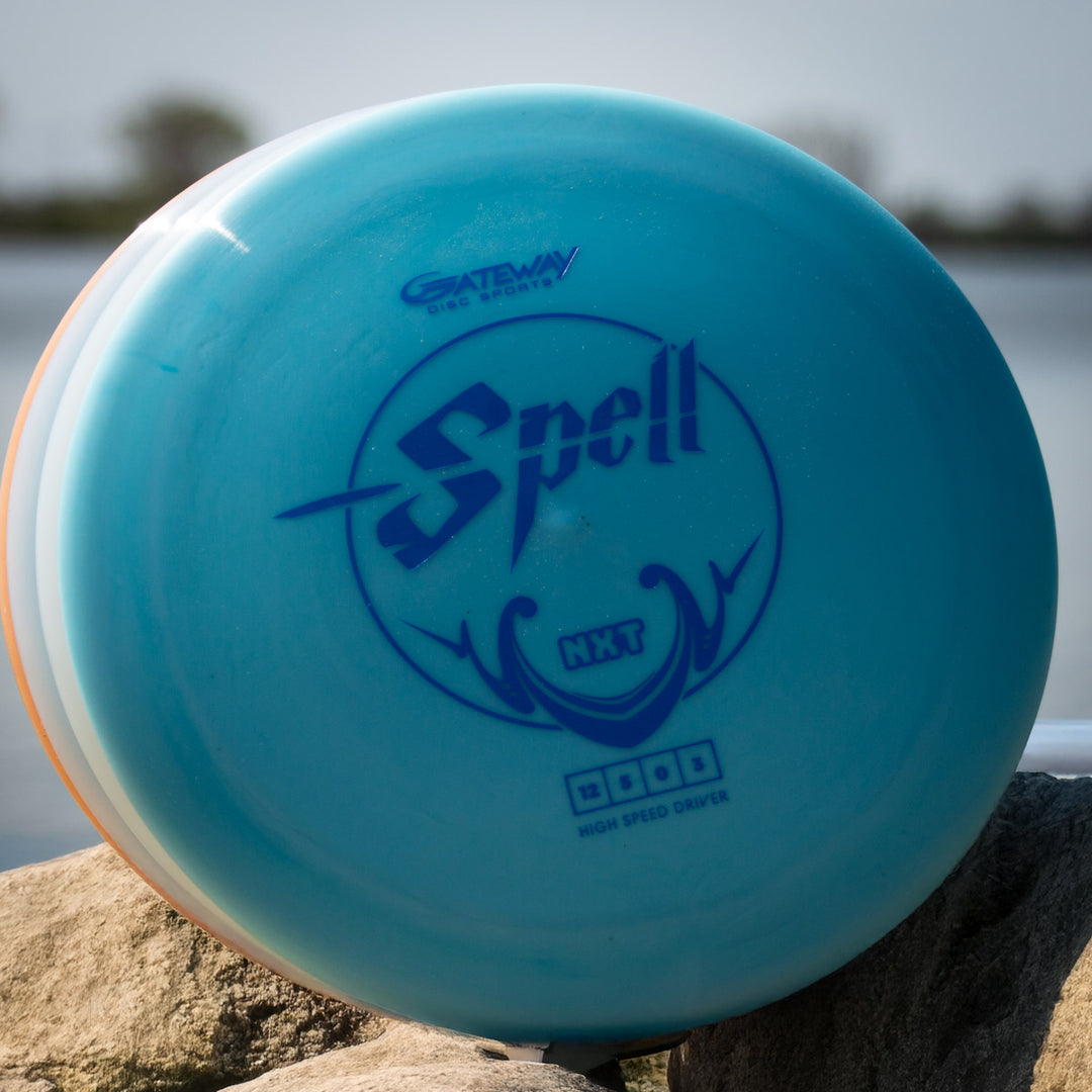A Spell - NXT overstable driver disc golf disc by Gateway, in blue, sits on rocks near water, poised to deliver powerful throws.