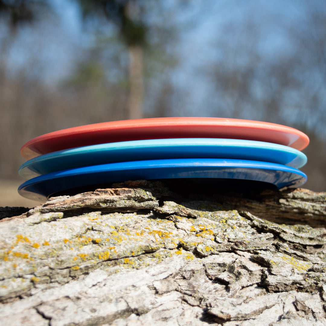 The Spell - Platinum disc golf discs, crafted from eco-friendly plastic in red and blue hues, are stacked on a tree trunk outdoors against a softly blurred natural backdrop.