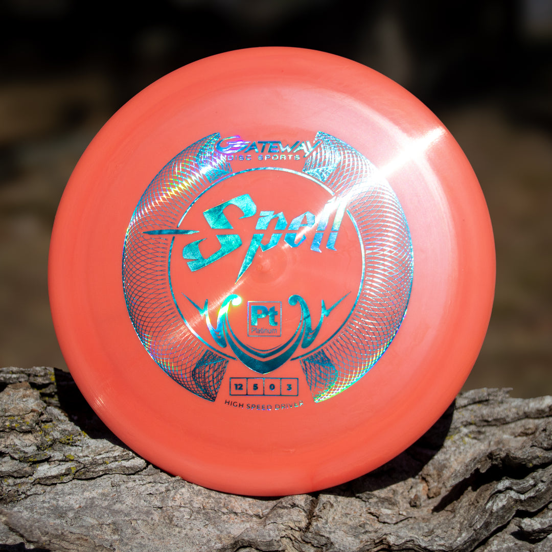 A Platinum Spell overstable driver by Gateway Disc Sports, featuring holographic text and design on eco-friendly plastic, rests outdoors on tree bark.