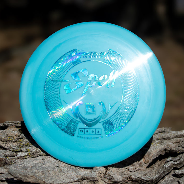 A light blue Spell - Platinum disc golf disc rests on rough tree bark, reflecting sunlight. This overstable driver is crafted from eco-friendly plastic for balanced performance and sustainability.