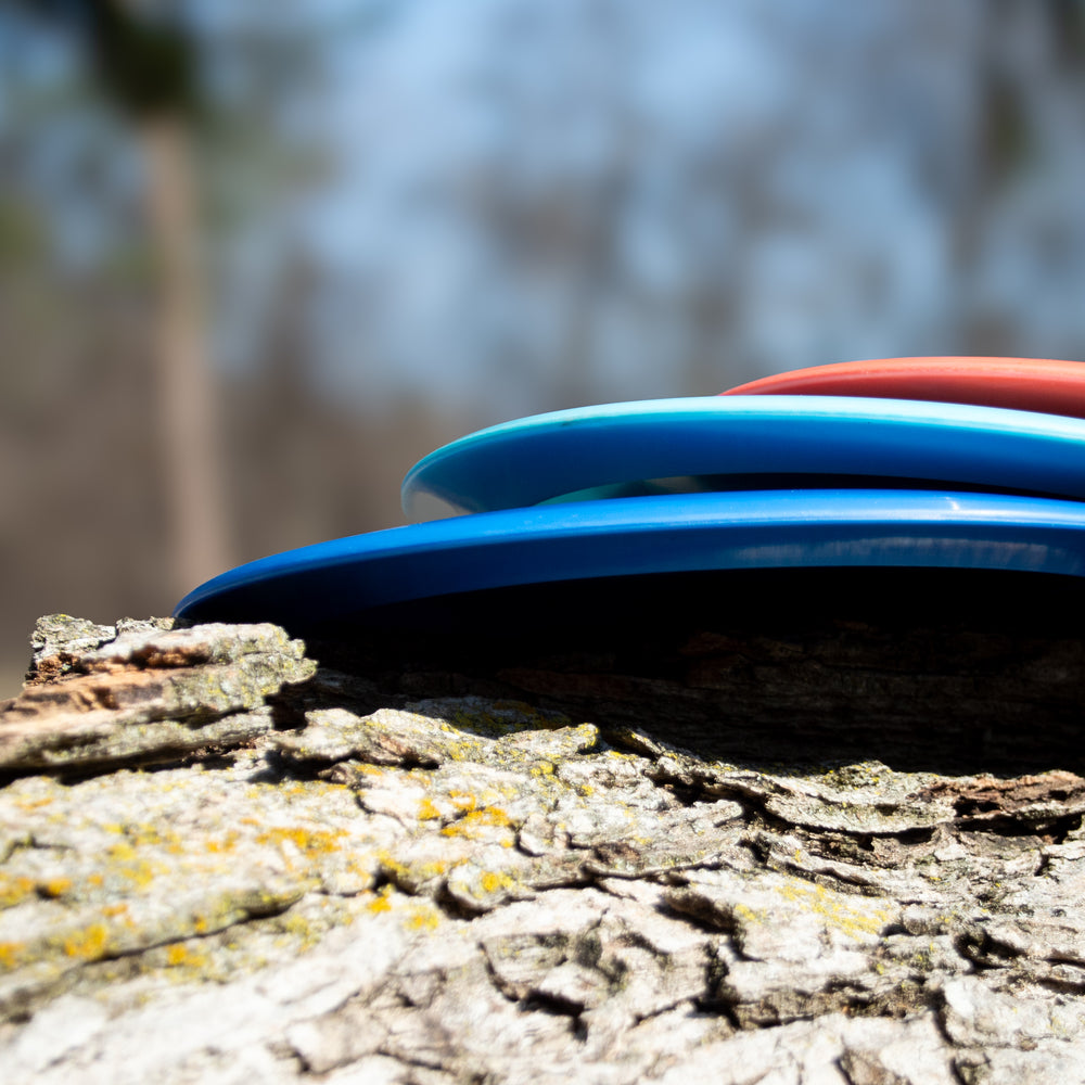 Three Spell - Platinum disc golf discs, crafted from eco-friendly plastic, are stacked side-on atop a rough tree trunk outdoors, with softly blurred trees and a blue sky in the background.
