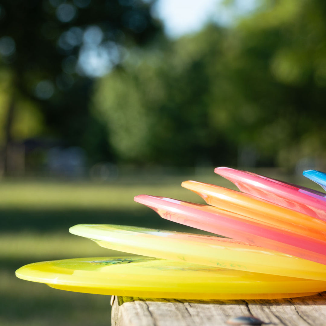 A stack of colorful plastic frisbees, including the Spirit - Diamond long distance driver, is arranged on a wooden surface outdoors with blurred green trees and grass in the background.