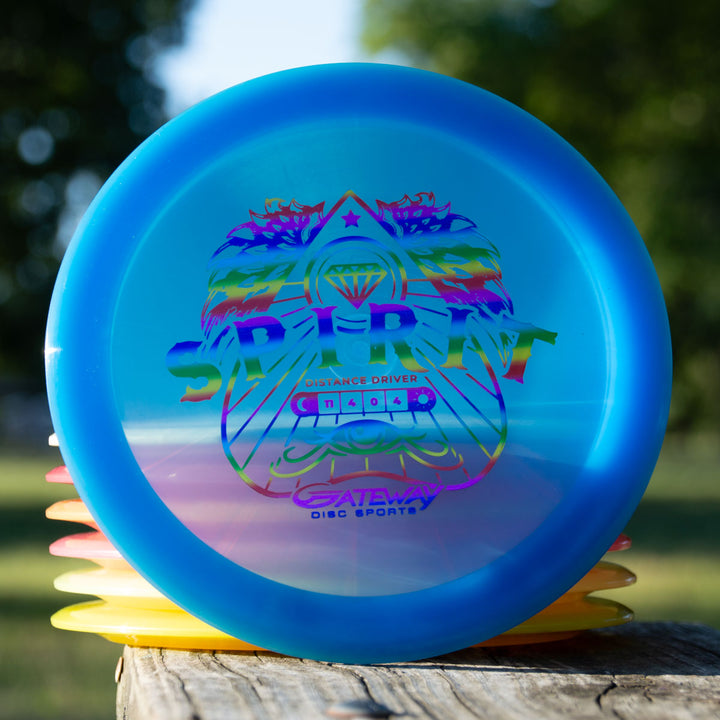 A stack of colorful long distance drivers sits on a wooden surface outdoors, with the top disc displaying a vibrant "Spirit - Diamond" logo—a high speed overstable driver perfect for powerful throws.