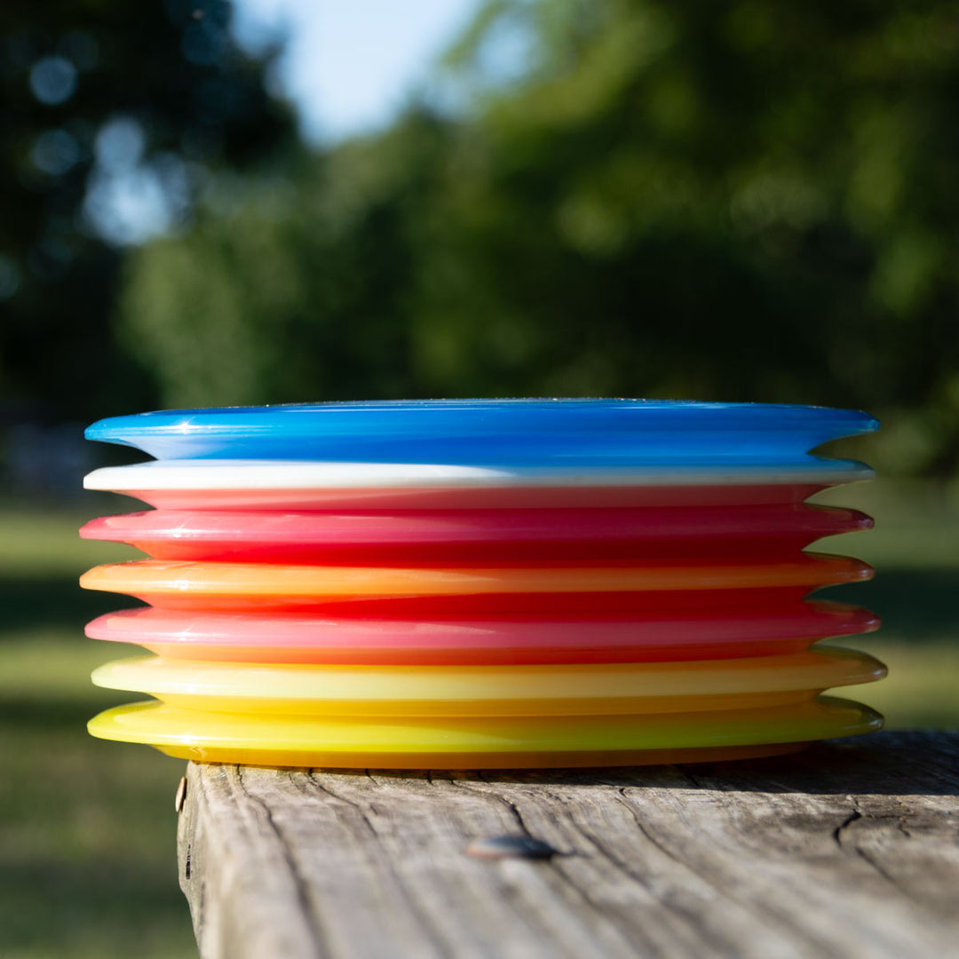 A stack of colorful plastic Spirit - Diamond flying discs, including a high speed overstable driver, rests on a wooden surface outdoors with blurred trees in the background.