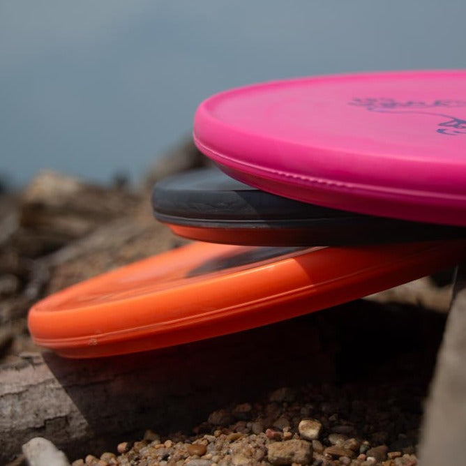 Three Reptilian DG Stego — Tar Pit discs in pink, black, and orange are stacked on a rocky surface outdoors, showcasing the rugged durability of Tar Pit plastic and the distinctive Stego design.