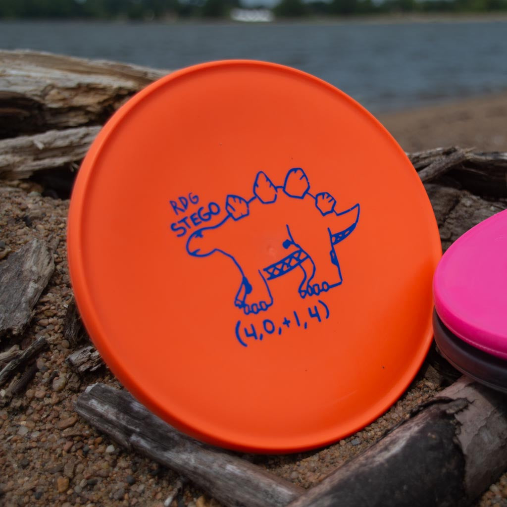 A Reptilian DG Stego — Tar Pit disc in orange, with a blue cartoon stegosaurus and "RDG STEGO (4,0,1,4)" text, is displayed on sand and driftwood near water.