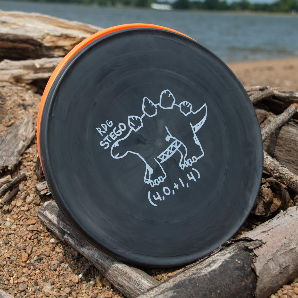A Reptilian DG Stego — Tar Pit disc, black and durable with a stegosaurus illustration and "RDG Stego (4.0, +1, 4)" label, sits on driftwood and sand beside the water.