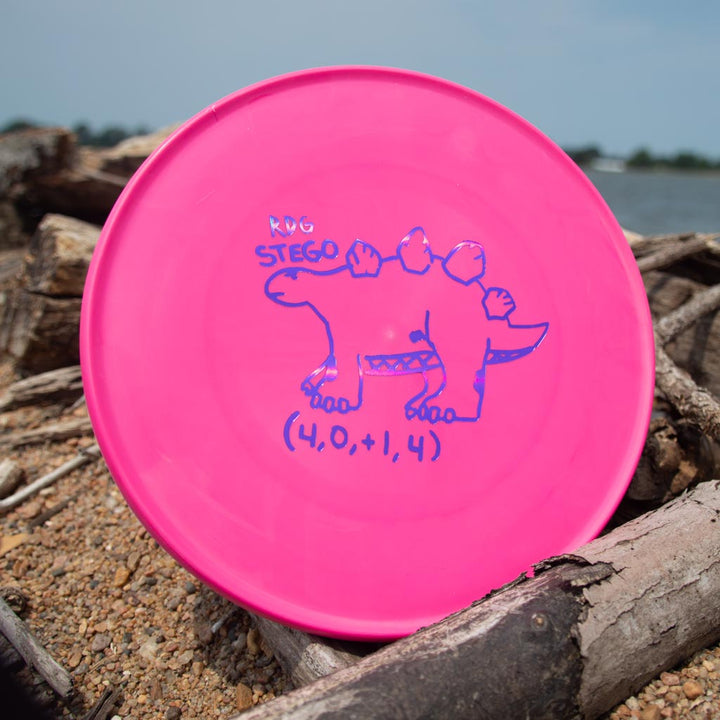 A Reptilian DG Stego — Tar Pit disc in pink 4S plastic, hand-drawn stegosaurus design and "RDG STEGO (4,0,+1,4)" label, rests on sand and driftwood near the water.