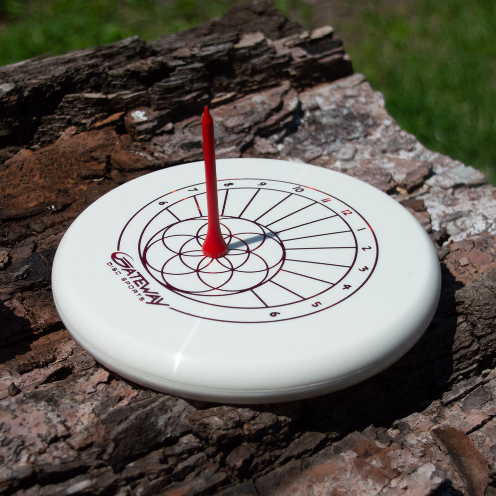 A "Sundial - Lil Wizard - Diamond" disc with a target design rests on tree bark, pierced vertically through the center by a red dart—evoking the image of a sundial struck at high noon.
