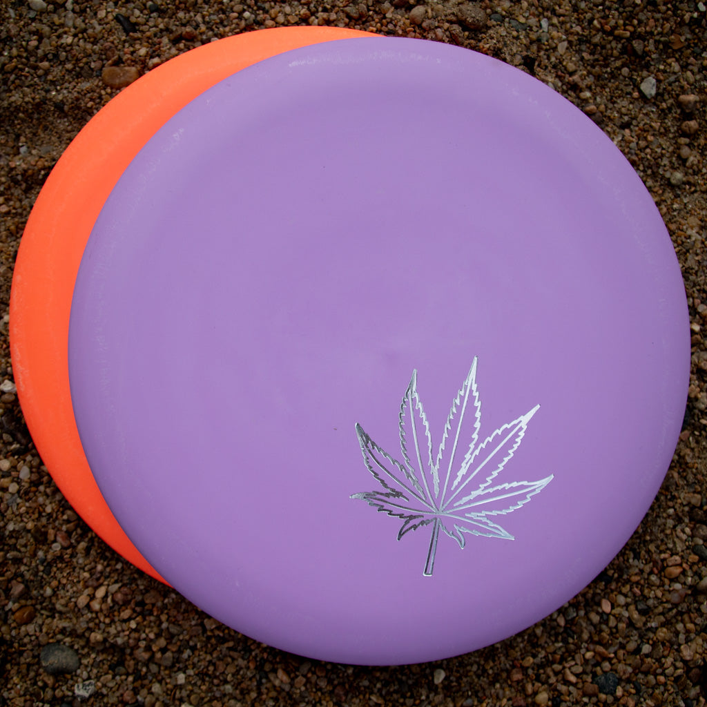 Two Wizard Leaf Suregrip Putters from Gateway Disc Sports rest on gravel: the top disc is purple with a silver iconic leaf, and the bottom disc is orange.