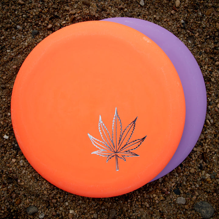 Two Wizard Leaf Suregrip Putters—one orange with the iconic leaf design and one purple—are stacked on sandy ground.