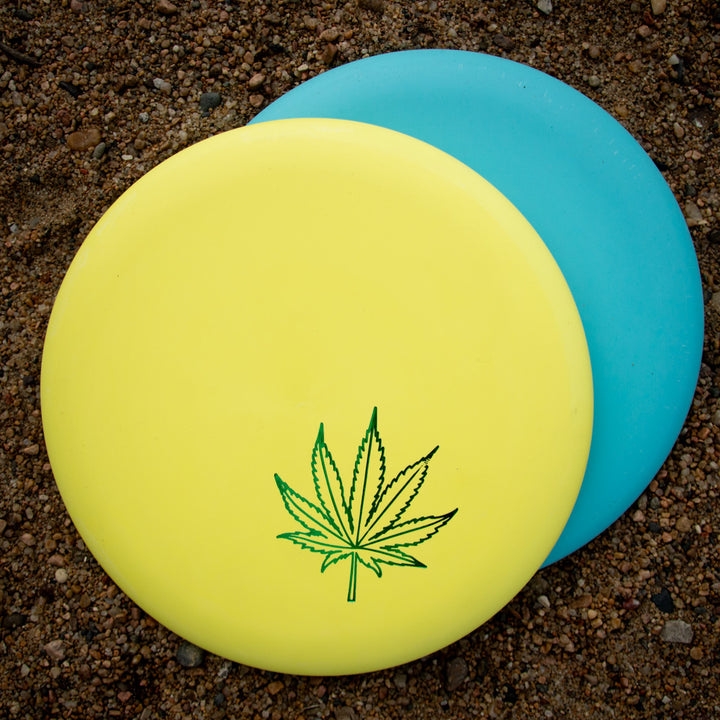 A yellow Wizard Leaf Suregrip Putter with an iconic leaf design overlaps a plain blue disc, both resting on gravelly ground.