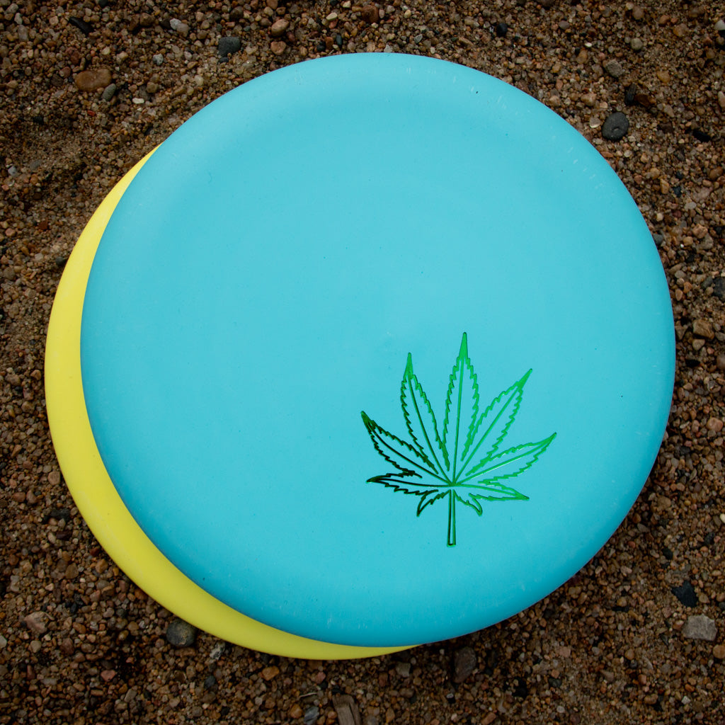 A blue Wizard Leaf Suregrip Putter from Gateway Disc Sports sits on a yellow putter atop gravelly ground.