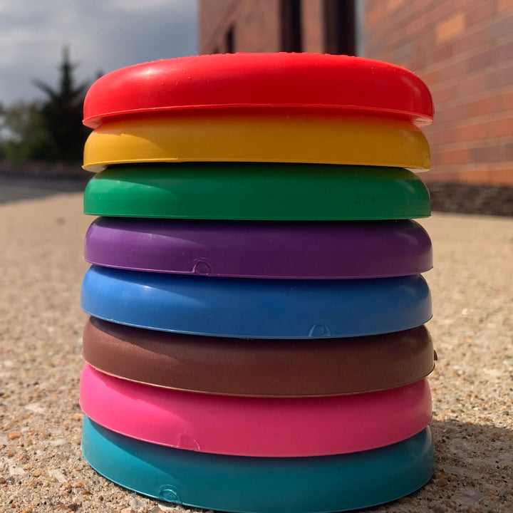 A stack of seven colorful Mini (Small) - SureGrip™ discs rests on a concrete surface outdoors near a brick building, ready for a fun game of disc golf.