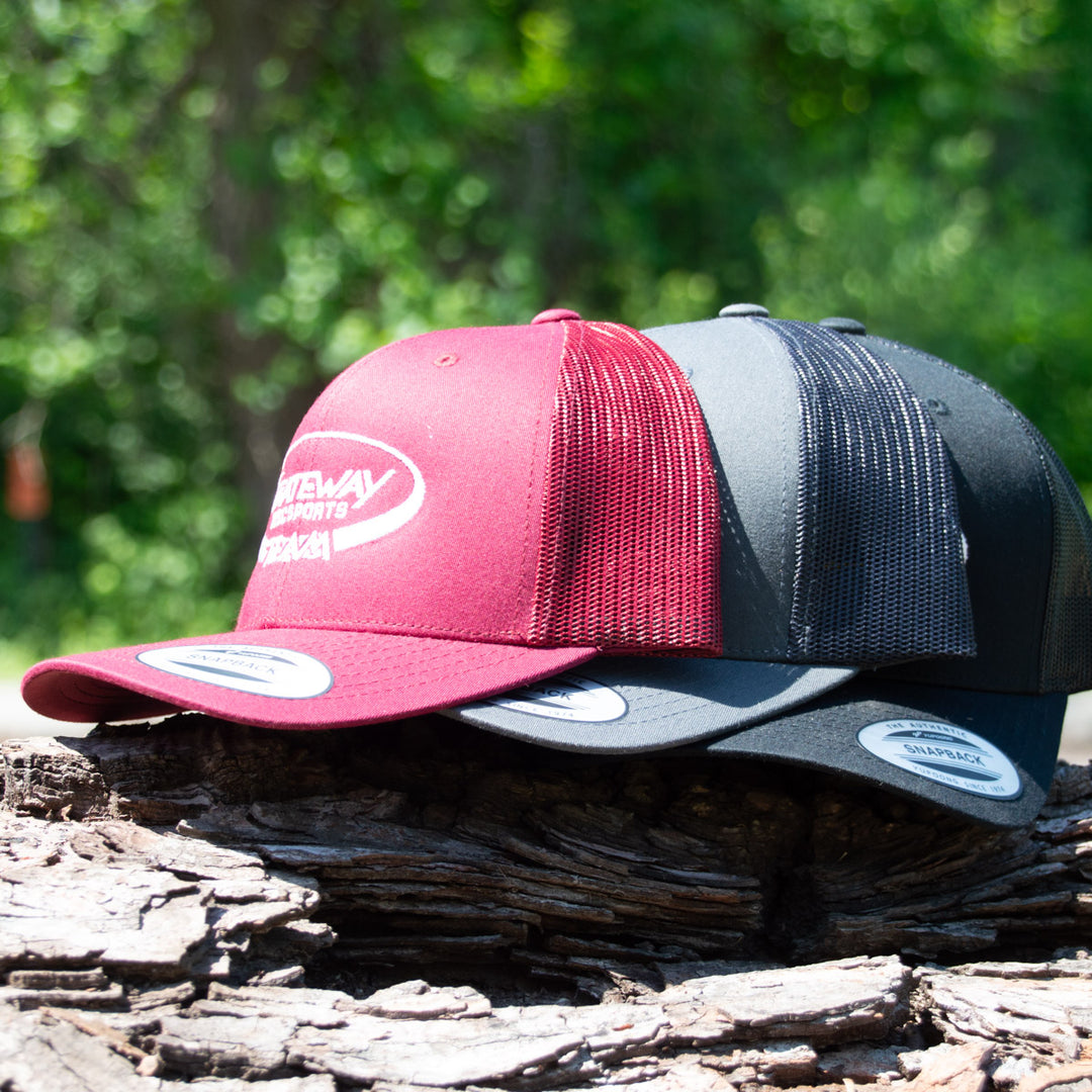 Two Team Gateway Snap Back hats, one red and one black, featuring white embroidered logos and snapback closures, are placed side by side on a tree log with greenery in the background.