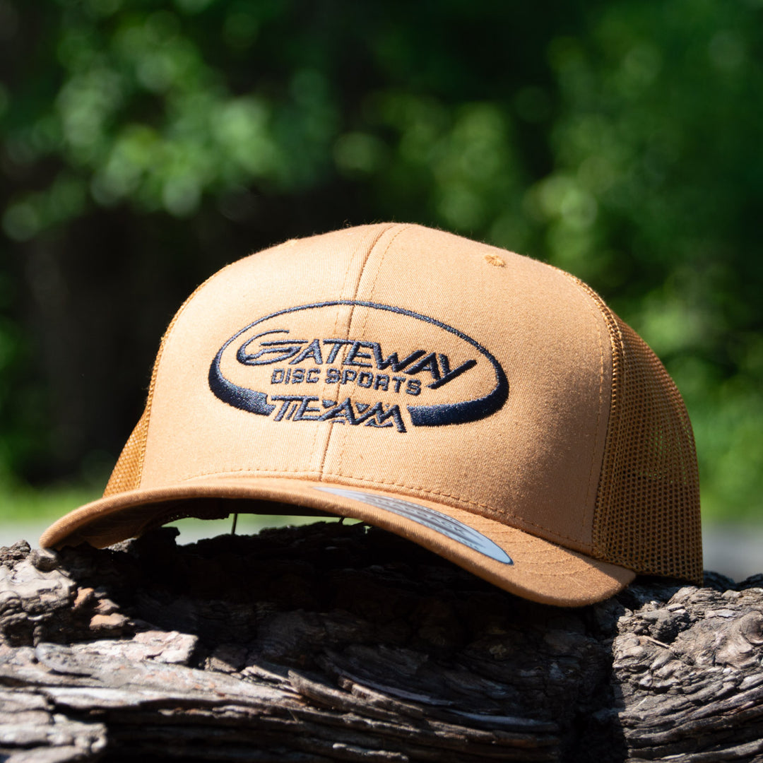 The Team Gateway Snap Back hat, a tan mesh back cap with a black "Gateway Disc Sports Team" logo, sits on a tree branch amidst lush greenery.