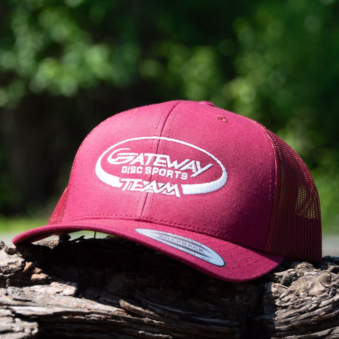 The Team Gateway Snap Back hat is a maroon polyester-cotton cap with a mesh back and "Gateway Disc Sports Team" embroidered in white, pictured outdoors on wood with greenery behind.
