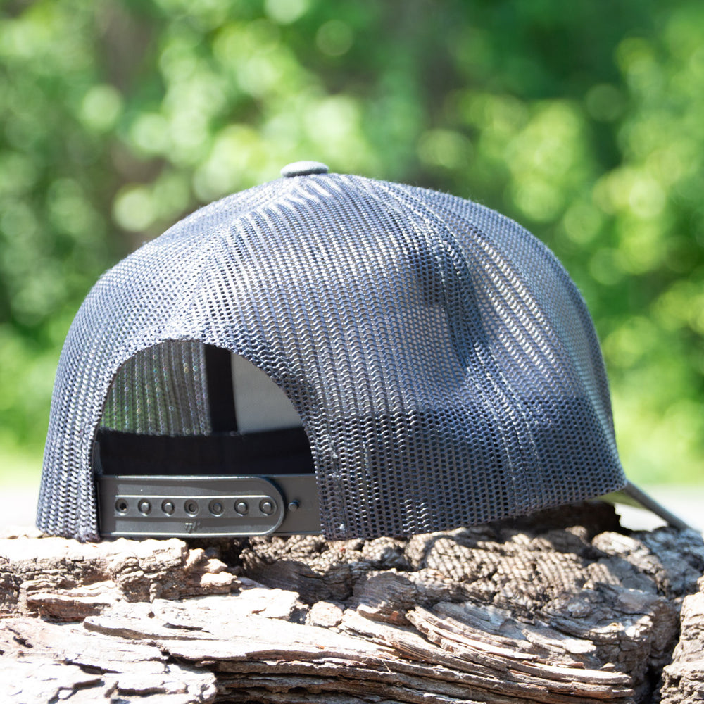 The Team Gateway Snap Back hat, featuring a gray mesh back, is displayed outdoors on a tree stump with greenery softly blurred in the background.