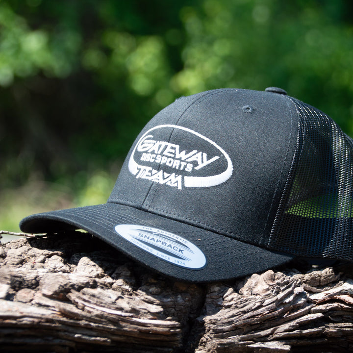 The Team Gateway Snap Back hat, a black poly-cotton cap with mesh back and snapback closure, features "Gateway Disc Sports Team" embroidered on the front and is displayed resting on a log amid blurred greenery.