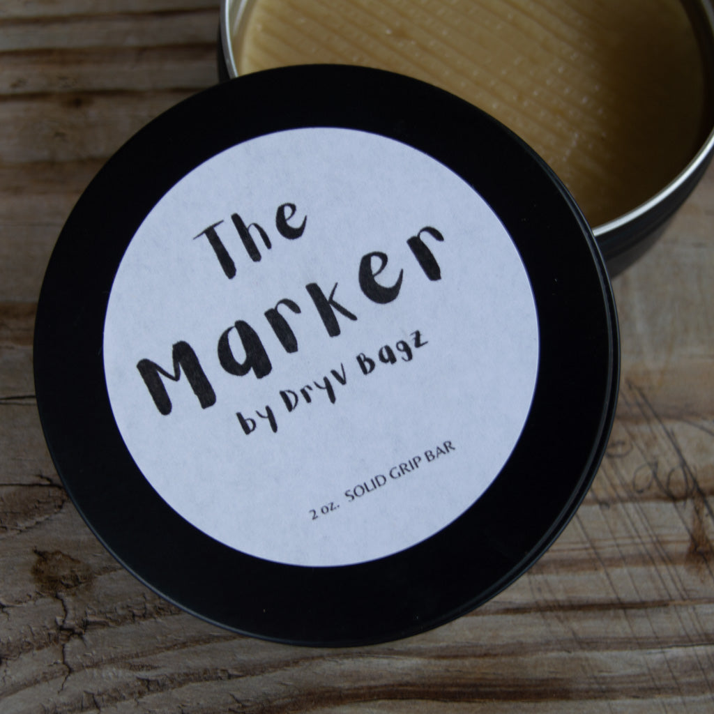 A round tin labeled "The Marker" sits on a wooden surface, its lid slightly open to reveal the solid 2 oz. grip enhancer bar inside.