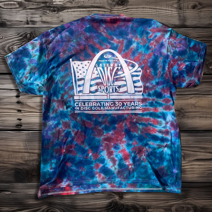 The 30th Anniversary - Tie Dyed T-Shirt features the "Gateway Disc Sports" anniversary logo with "Celebrating 30 Years in Disc Golf Manufacturing" on the back, shown on a wooden surface.