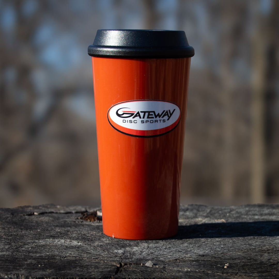 The Gateway 16oz Bronze Plastic Tumbler, featuring a black lid and "Gateway Disc Sports" logo, sits on a wooden surface outdoors—ideal for disc golf fans or as custom merch.