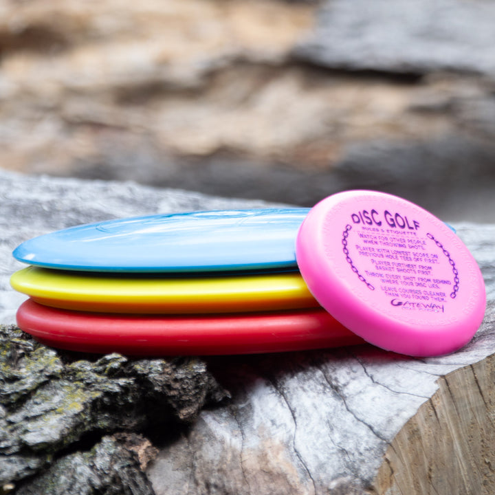 A 3-Disc Ultra Lightweight Starter Set with Mini rests on a weathered tree log amid rocks—the perfect beginner disc golf set for any outdoor adventure.