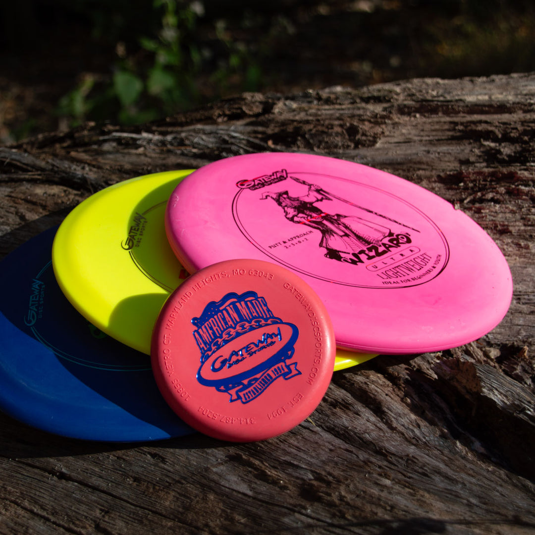 The 3-Disc Ultra Lightweight Starter Set with Mini, featuring pink, yellow, blue, and red discs including a putter, is stacked on weathered wood outdoors in sunlight with soft shadows.